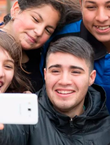 Un grupo de cinco jóvenes, hombres y mujeres, sonríen alegremente mientras se toman una selfie con un teléfono celular blanco. Se muestran cercanos y unidos en un ambiente exterior, transmitiendo una actitud positiva y de compañerismo.