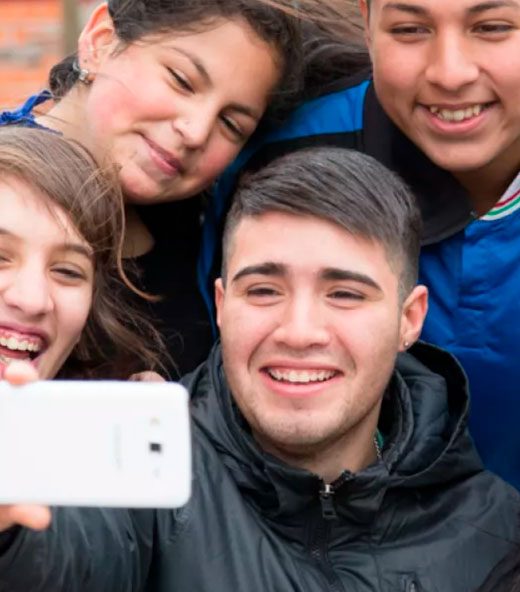 Un grupo de cinco jóvenes, hombres y mujeres, sonríen alegremente mientras se toman una selfie con un teléfono celular blanco. Se muestran cercanos y unidos en un ambiente exterior, transmitiendo una actitud positiva y de compañerismo.